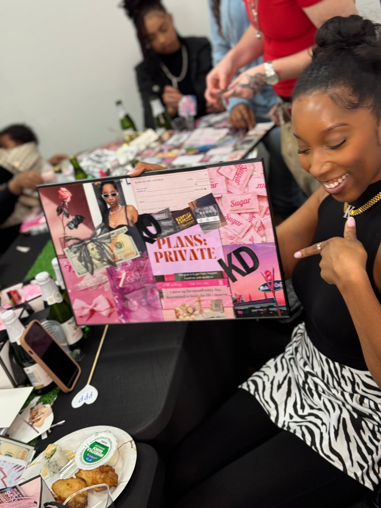 Woman holding a pink and black framed vision board with 'Plans: Private' text, surrounded by people and a table.